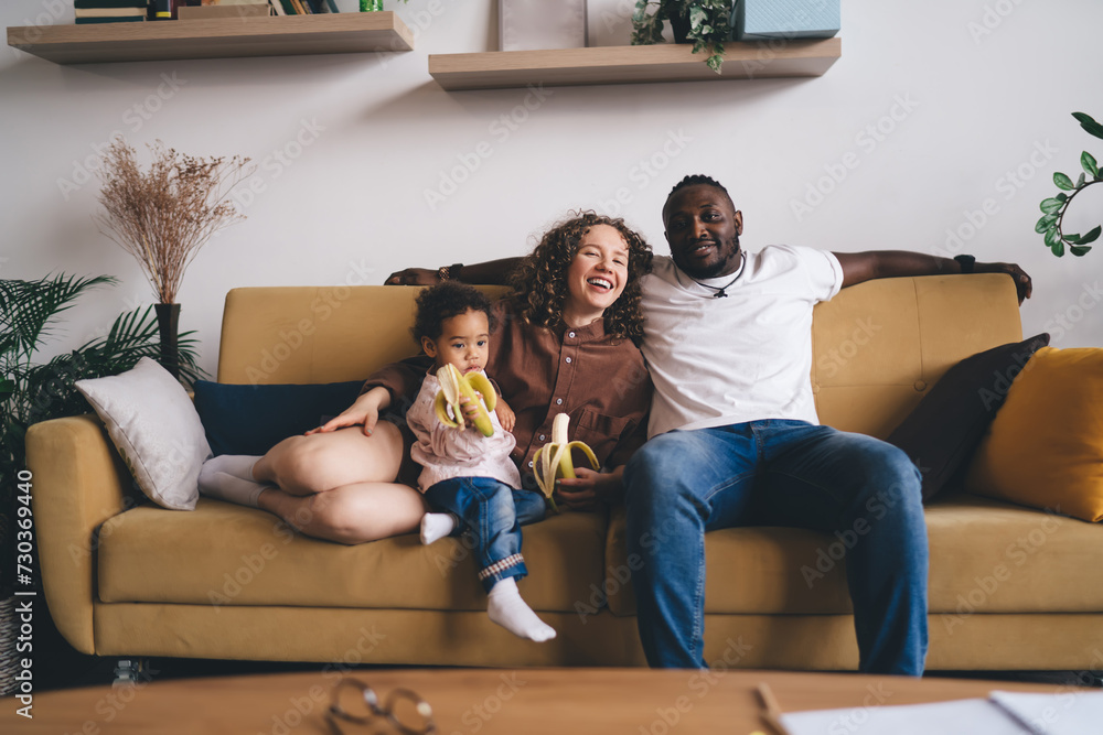 Obraz premium Cheerful diverse family eating banana while having fun together on sofa