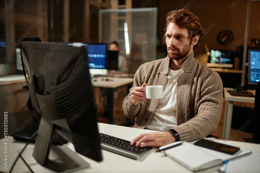 Portrait of bearded man using computer in office and drinking coffee while working late at night, copy space