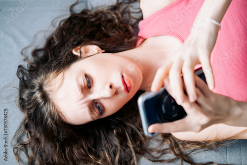 Young woman with long curly hair lies on the sofa and browses social networks on her phone with a serious look, top view