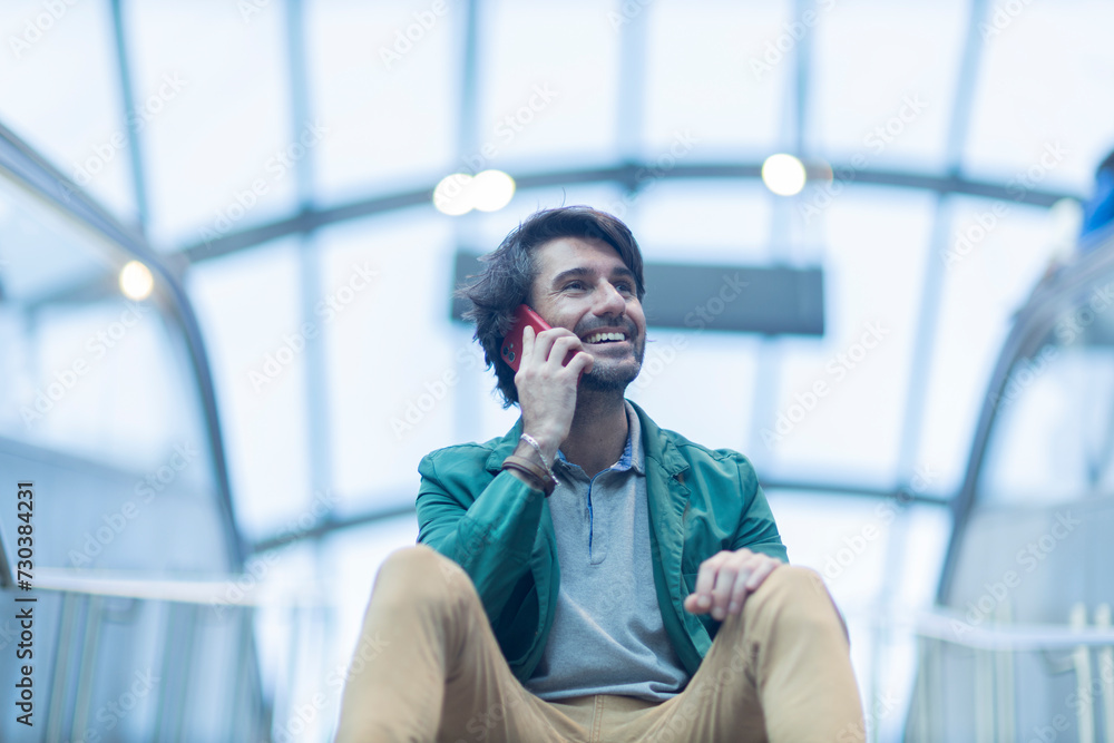 View of young man sitting down using a smartphone at indoor space with a blurred view landscape in the background. High quality photo. Texting on the phone. 