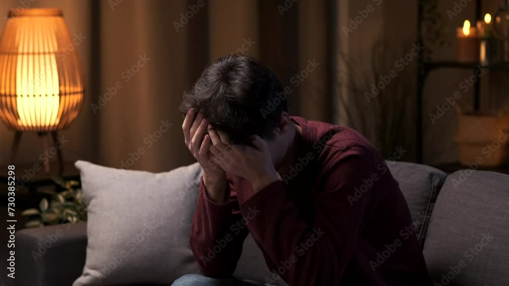 Caucasian man cries in sadness while sitting alone on the sofa in the living room in the evening. Negative emotions, crisis, mental health problems, troubles in private life.