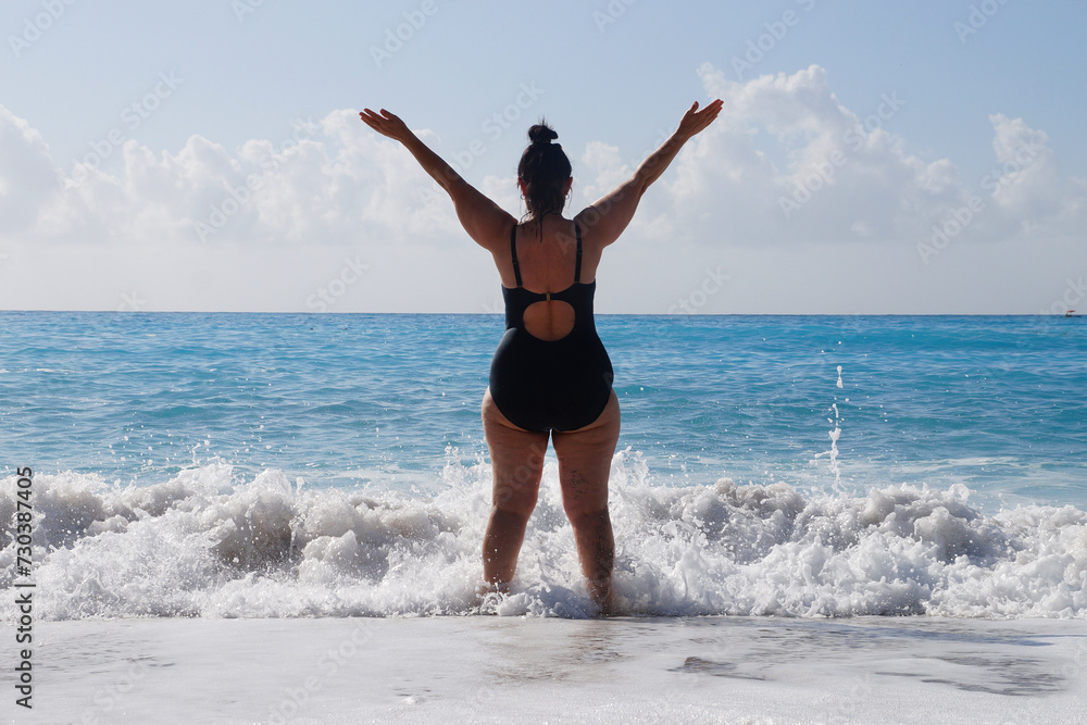 plump woman in a black swimsuit stands in the sea with her arms outstretched high, rear view plump woman in a black swimsuit stands in the sea with her arms outstretched high, rear view