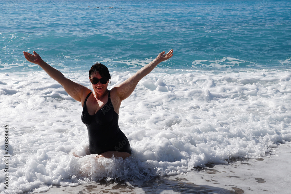 plump smiling woman in a black swimsuit and sunglasses sits in the sea with her arms stretched high plump smiling woman in a black swimsuit and sunglasses sits in the sea with her arms stretched high