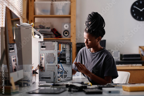 Black female technician sitting at table in workshop and repairing system unit using screwdriver