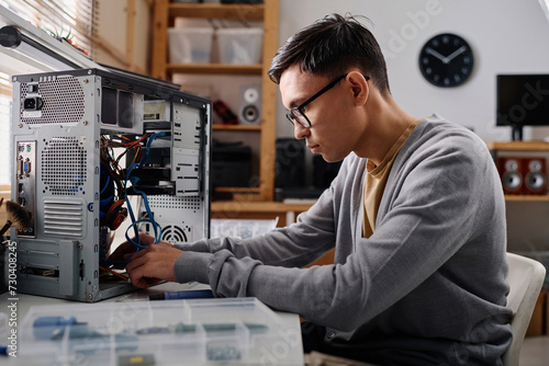 Young repairman arranging wires in system unit of computer sitting at desk in modern workshop