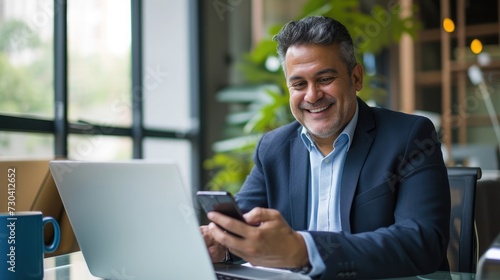 Middle aged Hispanic business manager ceo using cell phone mobile app, laptop. Smiling Latin or Indian mature man businessman holding smartphone sit © buraratn