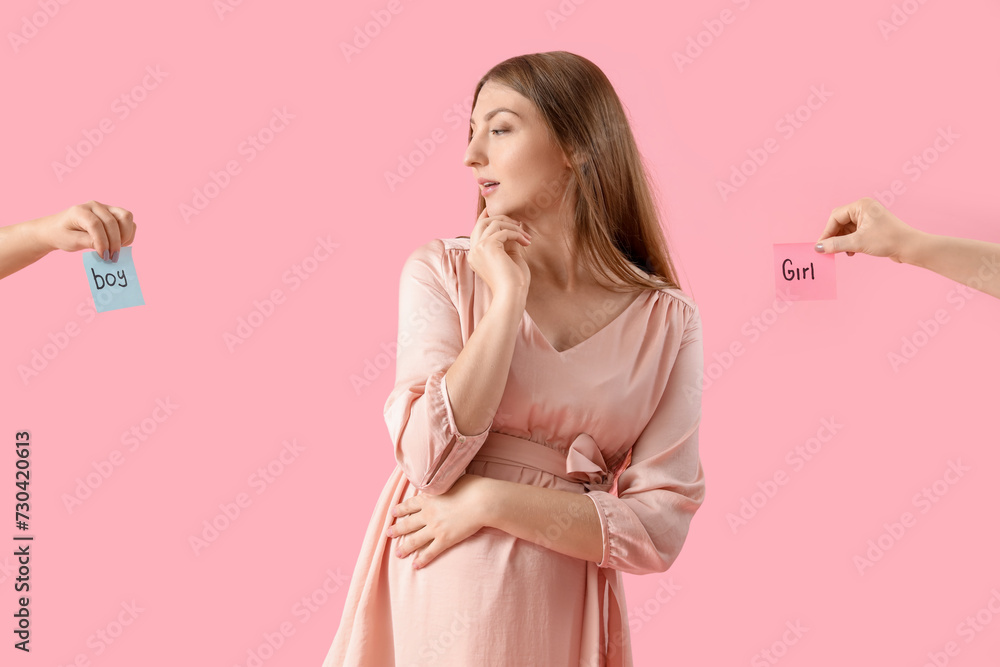 Fototapeta premium Thoughtful young pregnant woman with words BOY and GIRL on papers against pink background