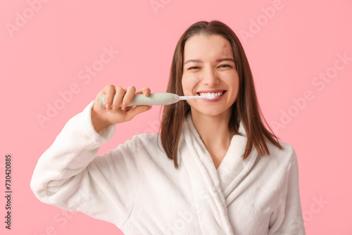 Beautiful woman in bathrobe with electric toothbrush on pink background
