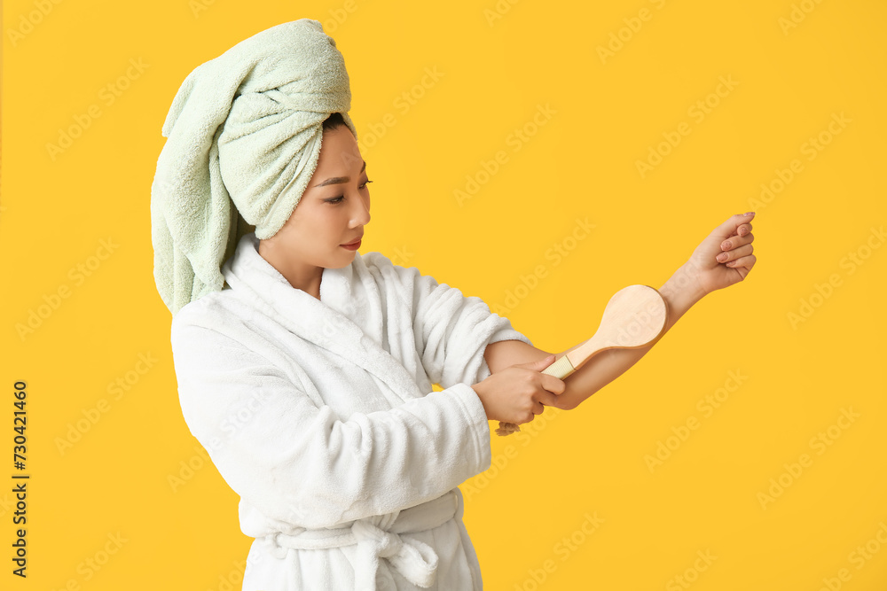 Young Asian woman in bathrobe with massage brush after shower on yellow background