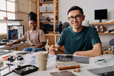 Caucasian technician smiling at camera while soldering cellphone, his black coworker sitting on background