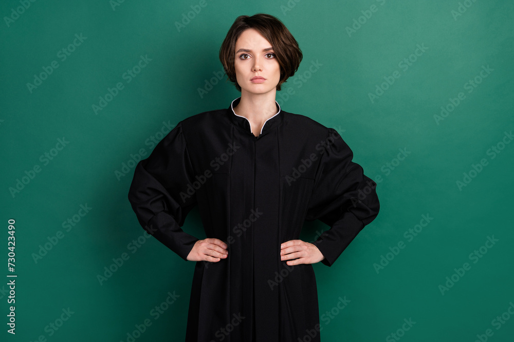 Photo of serious beautiful judge woman attorney hold hands waist wear black robe isolated on green color background Photo of serious beautiful judge woman attorney hold hands waist wear black robe isolated on green color background