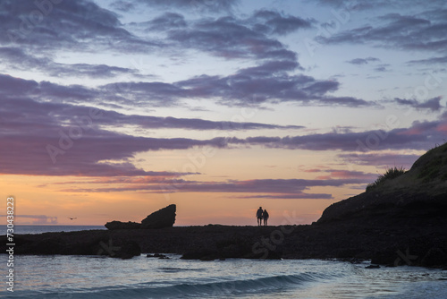 Couple standing a rocky headland at sunset watchng the sun go down. Spirits Bay, Northland, New Zealand. photo