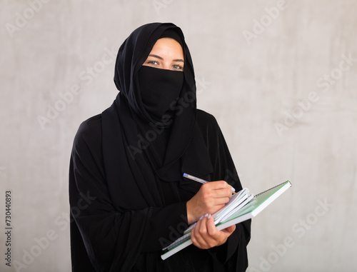 young calm girl with covered face by black burka holds several notebooks in her hands and writes important note in her diary