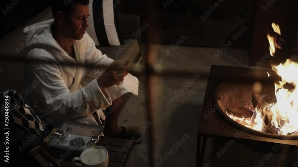Man in white bathrobe sits with cigar and cup of coffee on table in front of portable fireplace. Slow motion Man in white bathrobe sits with cigar and cup of coffee on table in front of portable fireplace. Slow motion
