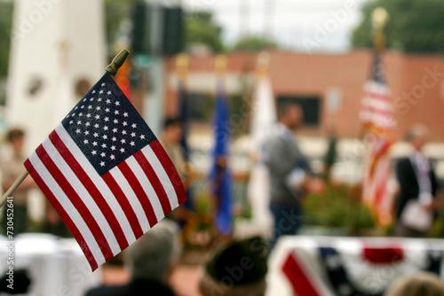 Horizontal close-up of a US Flag with blurred Memorial Day ceremony in the Background.