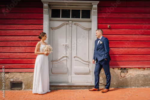 Valmiera, Latvia - July 7, 2023 - Bride and groom standing apart, facing each other smiling, in front of a red wooden building with a white door