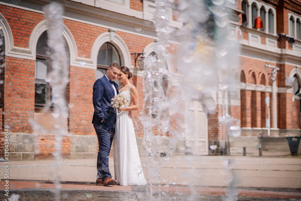 Valmiera, Latvia - July 7, 2023 - Bride and groom by a fountain, brick building in background, groom in blue suit, bride in white gown with bouquet