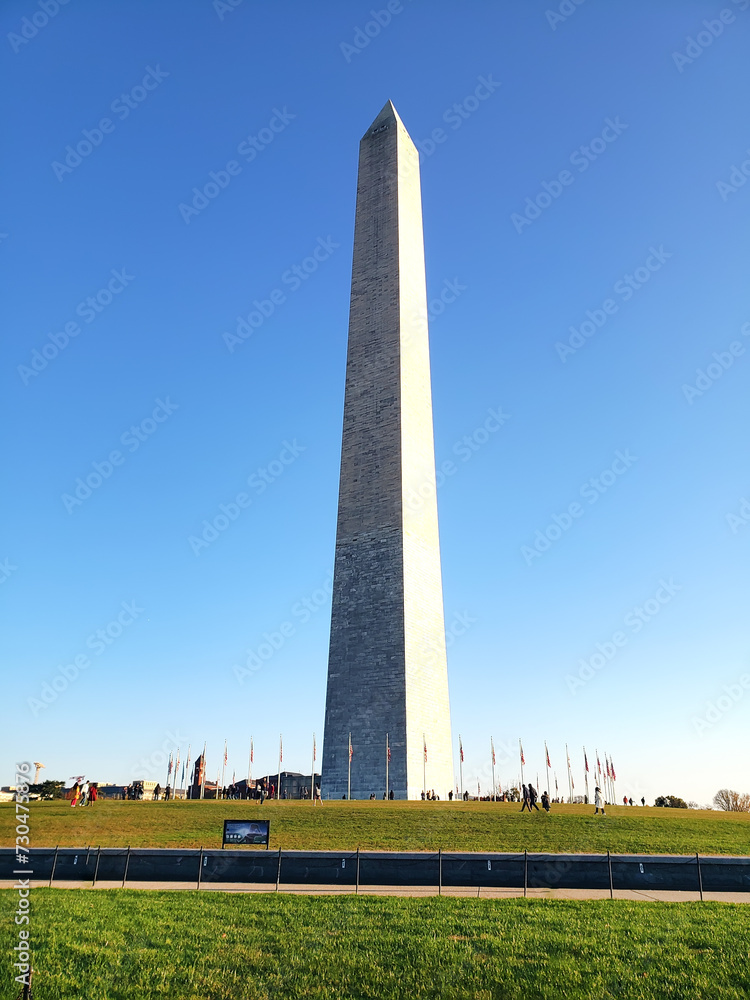 Washington Monument in Washington DC in clear blue sky day. People walk by the pond.