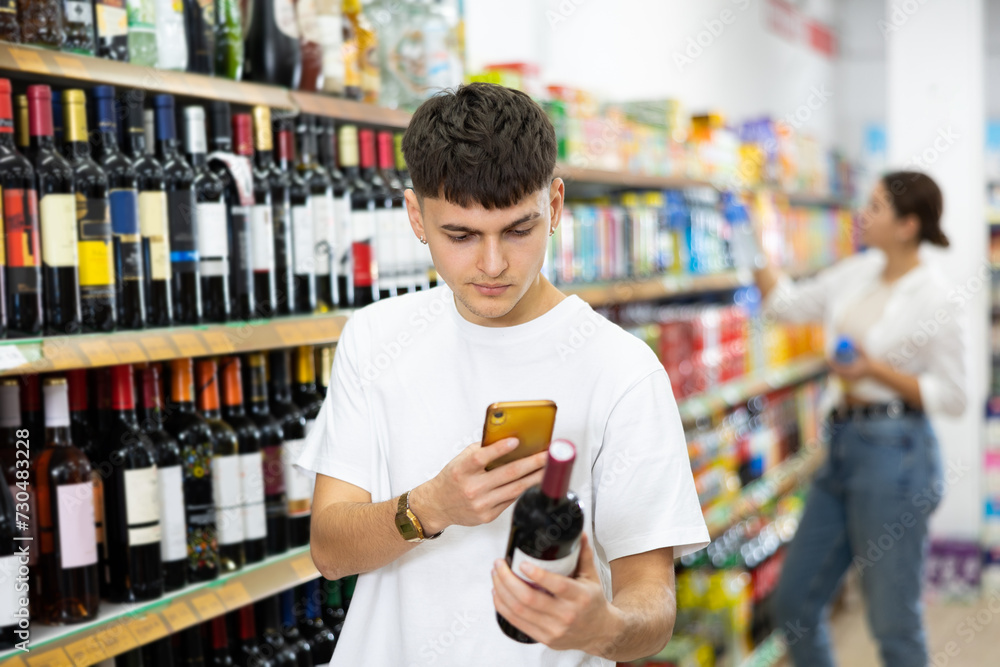 Focused interested young guy making purchases in supermarket, using smartphone to scan barcode on bottle of wine. Modern shopping concept