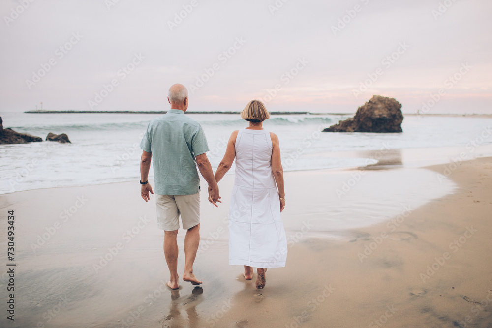 elderly couple holding hands and walking away from the camera at the beach in a dress and shorts at sunset