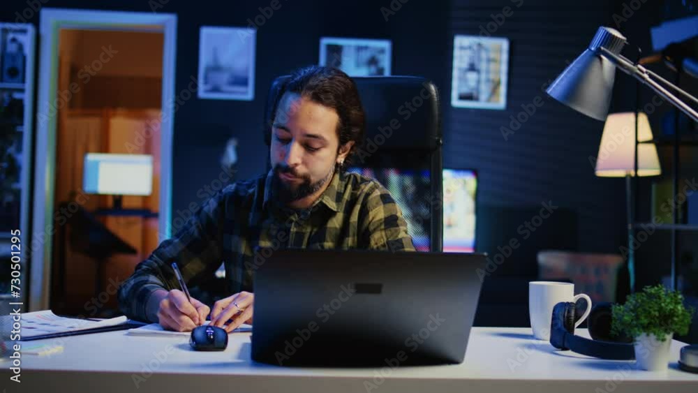 Remote employee working at home office desk, reading information on laptop and writing in notebook. Freelancer noting down ideas on paper using pen while teleworking, camera B Remote employee working at home office desk, reading information on laptop and writing in notebook. Freelancer noting down ideas on paper using pen while teleworking, camera B