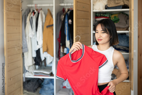Glad young Asian woman looking at dresses held in hand for choice in dressing room photo