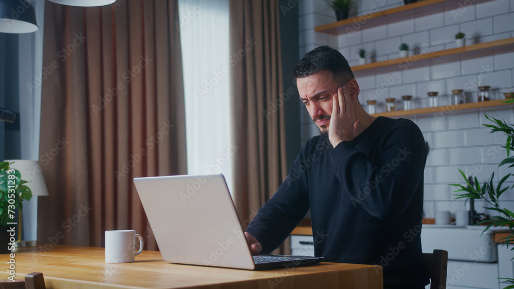 Tired man having pain from a headache or migraine rubbing his head while using laptop in the kitchen at home Tired man having pain from a headache or migraine rubbing his head while using laptop in the kitchen at home
