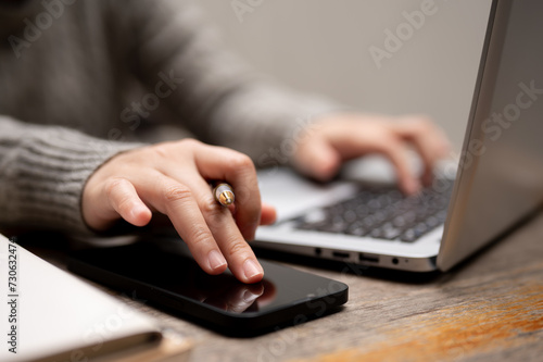 A woman using her smartphone while typing on a keyboard, working on her laptop at a table.