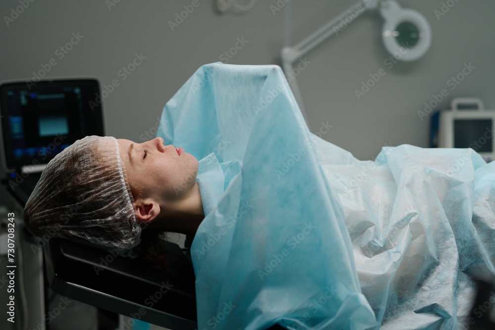 Young female patient in scrubs cap lying in front of camera in operating room against computer screen before medical procedure Young female patient in scrubs cap lying in front of camera in operating room against computer screen before medical procedure