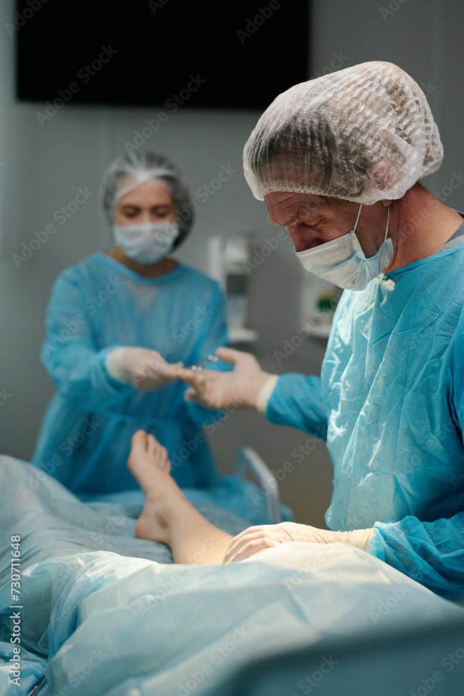 Mature surgeon taking medical tool from hand of female assistant in medical scrubs, gloves, cap and mask during operation Mature surgeon taking medical tool from hand of female assistant in medical scrubs, gloves, cap and mask during operation
