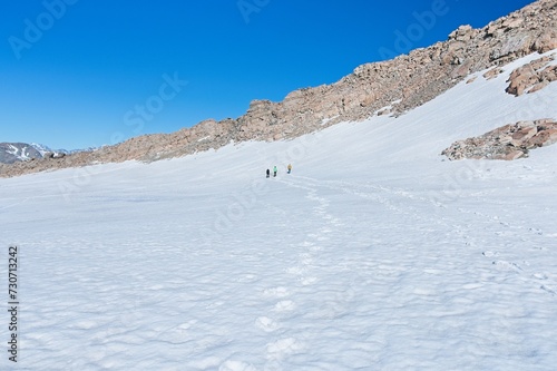 Adventure Seekers in Snowy Wilderness: Three Figures Hiking Amidst Majestic Rocky Mountains Under a Clear Blue Sky.