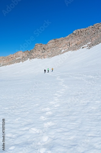 Adventure Seekers in Snowy Wilderness: Three Figures Hiking Amidst Majestic Rocky Mountains Under a Clear Blue Sky.