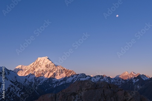 Scenic Sunrise at Mount Cook: Snowy Mountain Peaks Glowing in Golden Light, Accompanied by the Moon in New Zealand's Breathtaking Landscape