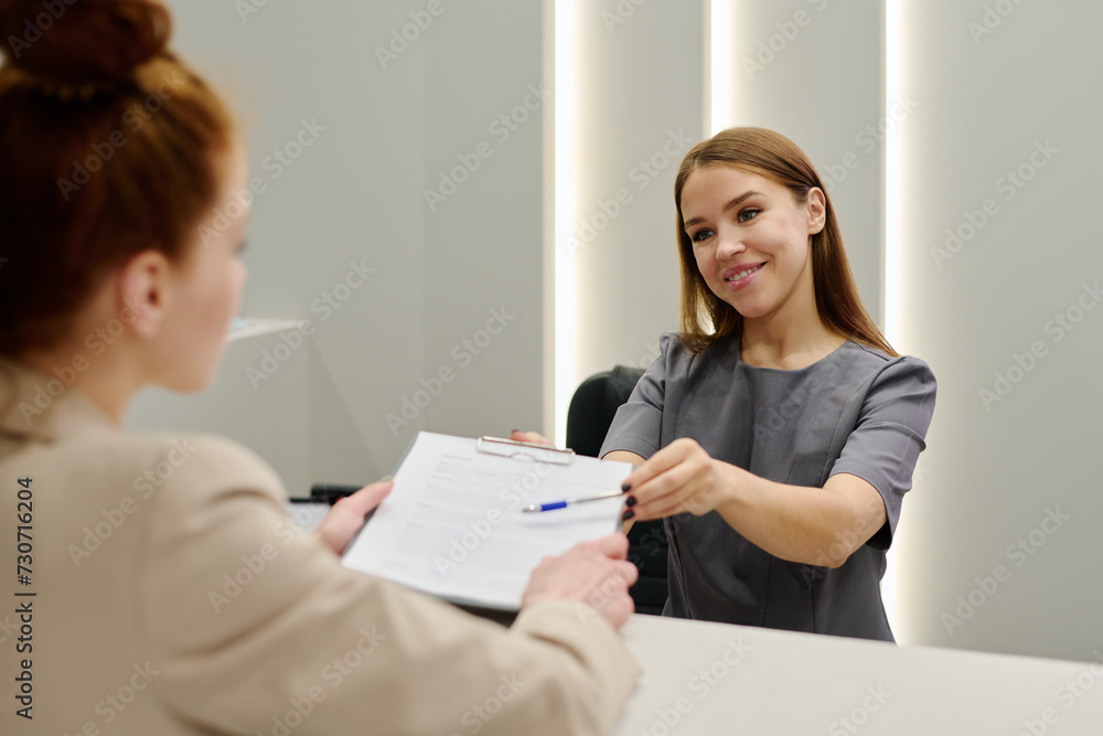 Happy young receptionist or clinician in uniform passing medical document to female patient standing in front of counter Happy young receptionist or clinician in uniform passing medical document to female patient standing in front of counter
