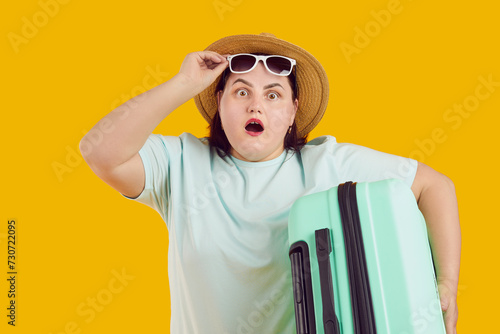 Portrait of shocked surprised fat woman with open mouth in straw hat holding suitcase raising sunglasses on studio yellow background. Funny tourist is going on summer holiday trip. Vacation concept.