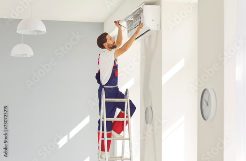 Service worker stands on ladder in modern flat or office, sets up new AC, repairs some parts, or removes dirty purifier. Air conditioning installation, full warranty repair and maintenance concept