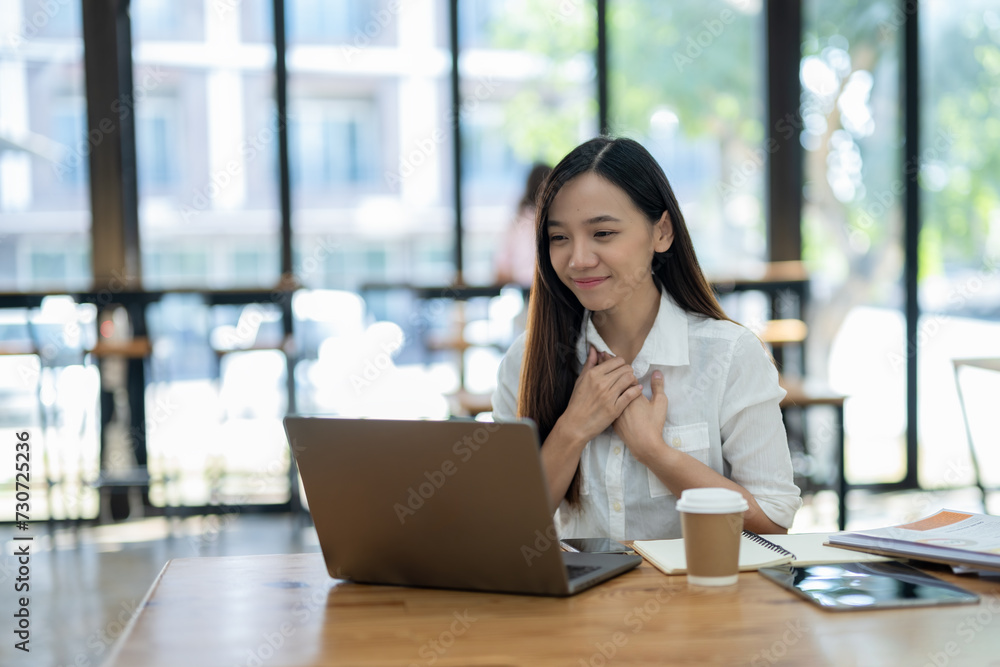 Content Asian businesswoman smiling and feeling grateful during a pleasant conversation over a laptop in a sunny office..