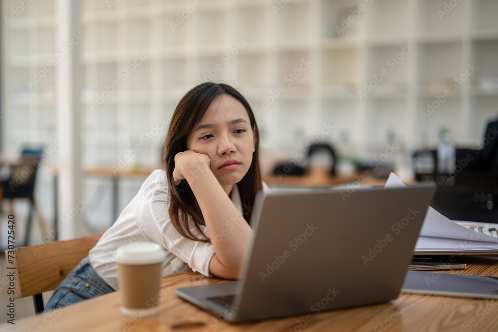 A stressed young Asian businesswoman massages temples while focusing on a laptop screen in a well-lit office environment.. A stressed young Asian businesswoman massages temples while focusing on a laptop screen in a well-lit office environment..
