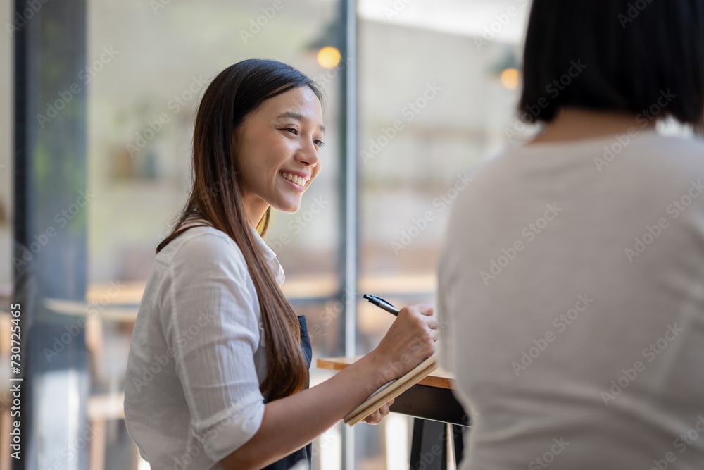 Fototapeta premium A cheerful Asian cafe owner takes an order from a customer, writing on a notepad with a warm and welcoming smile..
