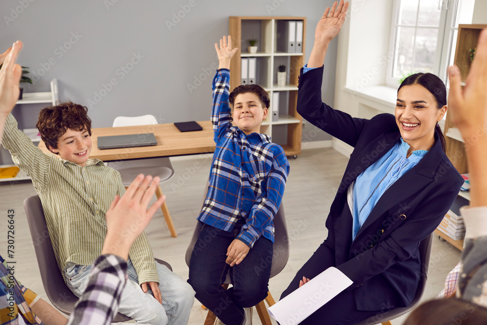 Obraz premium Group of junior school students and teacher raising hands together while sitting in circle in classroom. Happy smiling boys and girls classmates and their teacher having fun together during lesson