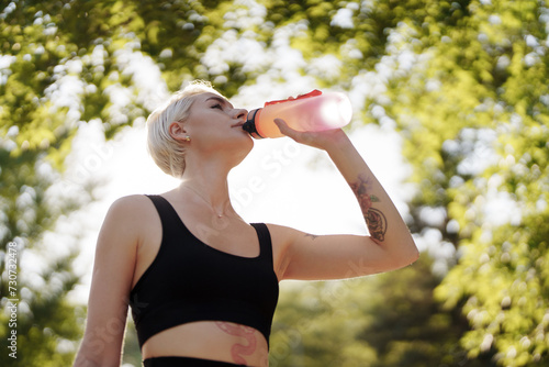 Backlit by sunlight, a woman pauses to drink water during her morning workout in the park