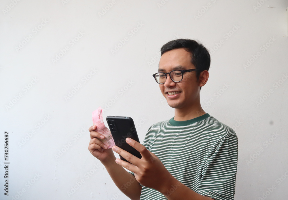 Adult Asian man with glasses showing happy face expression while holding mobile phone and paper money, isolated background Adult Asian man with glasses showing happy face expression while holding mobile phone and paper money, isolated background