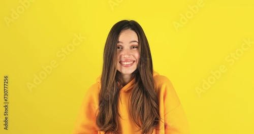 Shocked smiling shy charming confused young woman in yellow hoody posing isolated on yellow background studio. Girl looking at camera smile cover mouth with hand say oops. photo