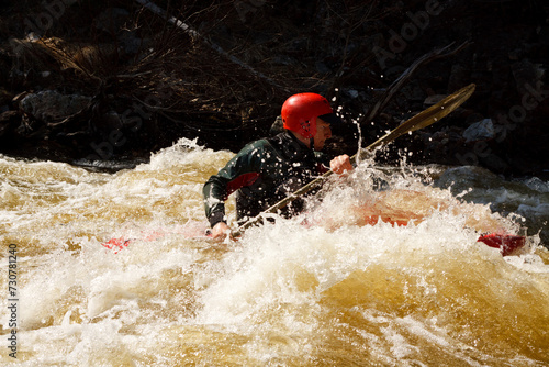 Man in a red helmet kayaking in a river with white water and rocks in the background. photo
