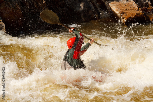 Man in a red helmet paddles a kayak down a river with whitewater rapids.