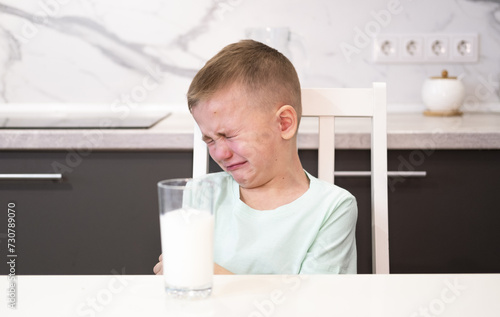 Fotografija An unhappy child refuses to drink milk for breakfast and sits in kitchen without an appetite