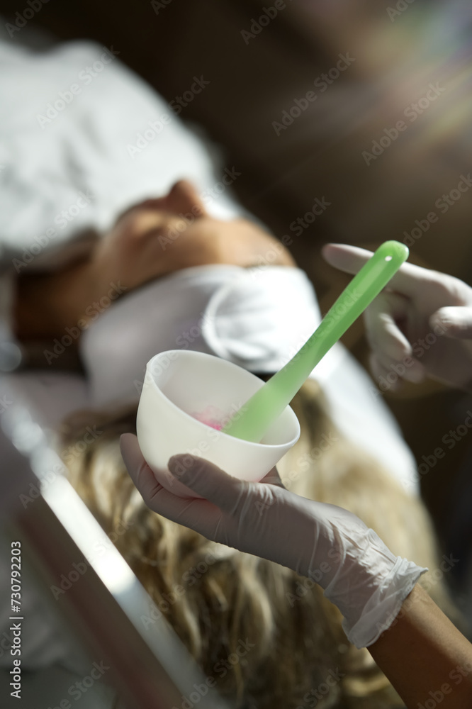 application of a cosmetic face mask in a bowl, woman in the background lying on a treatment couch