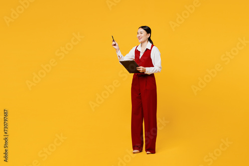 Full body young employee business woman of Asian ethnicity wears red vest shirt work at office hold clipboard paper account documents point aside isolated on plain yellow background. Career concept. photo