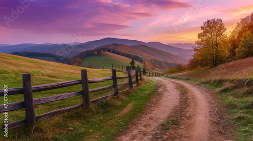 Rural dirt road  wooden fence  green hills in mountain valley at sunset in autumn. Landscape with country road  meadows  trees  purple sky with pink clouds at twilight