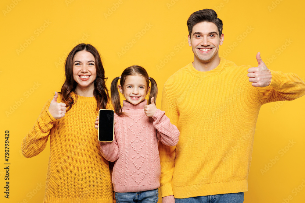 Young parents mom dad with child kid girl 7-8 years old wear pink sweater casual clothes use blank screen area mobile cell phone show thumb up isolated on plain yellow background. Family day concept. Young parents mom dad with child kid girl 7-8 years old wear pink sweater casual clothes use blank screen area mobile cell phone show thumb up isolated on plain yellow background. Family day concept.